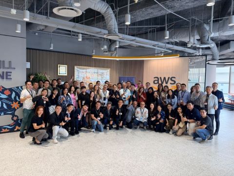 Large group of attendees posing indoors with thumbs up in front of a wall bearing the AWS logo