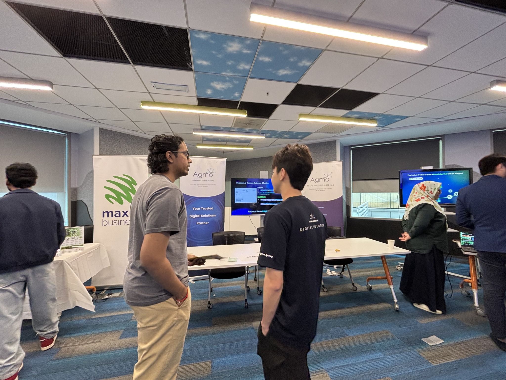 Two attendees conversing in front of Agmo banners and a screen at the Maxis AI First event