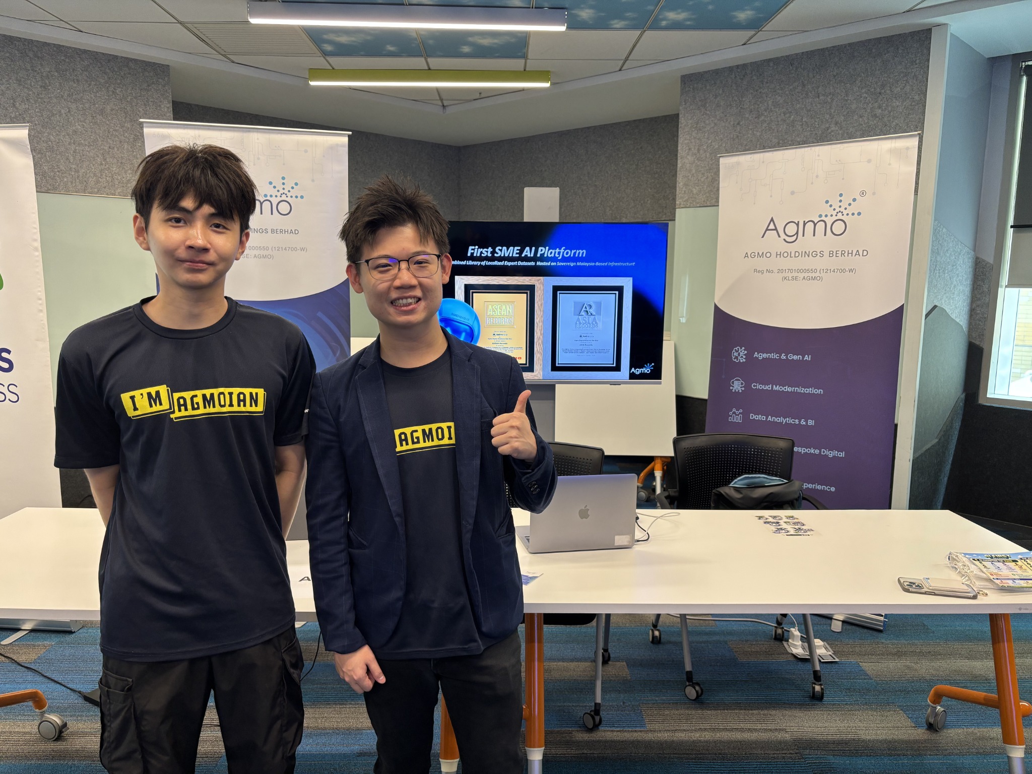 Two Agmo team members in branded t-shirts standing beside an Agmo screen display