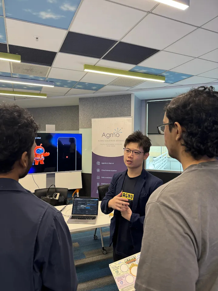 An Agmo speaker explaining a demo to two attendees in front of a screen and Agmo banner
