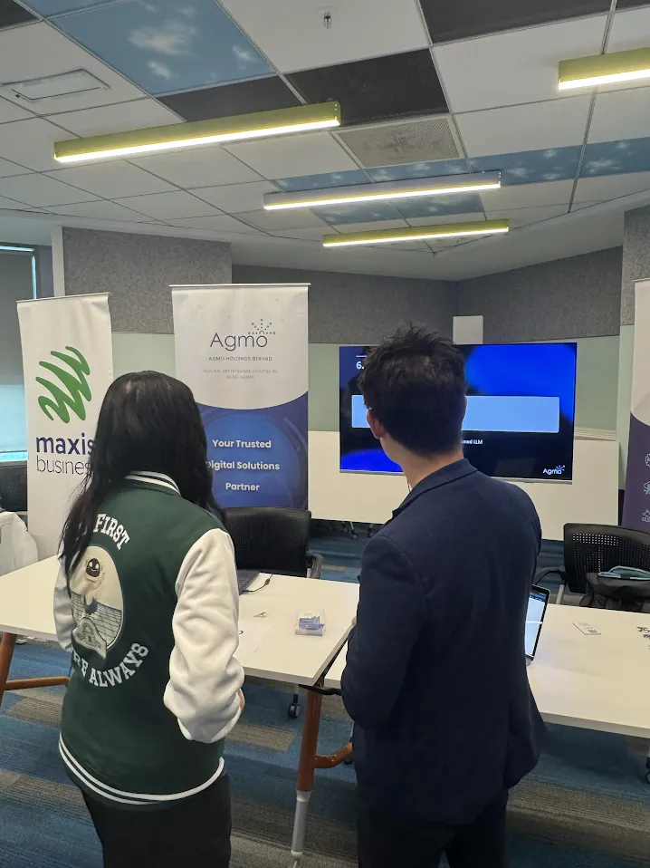Two attendees viewing a presentation on screen beside Maxis and Agmo banners